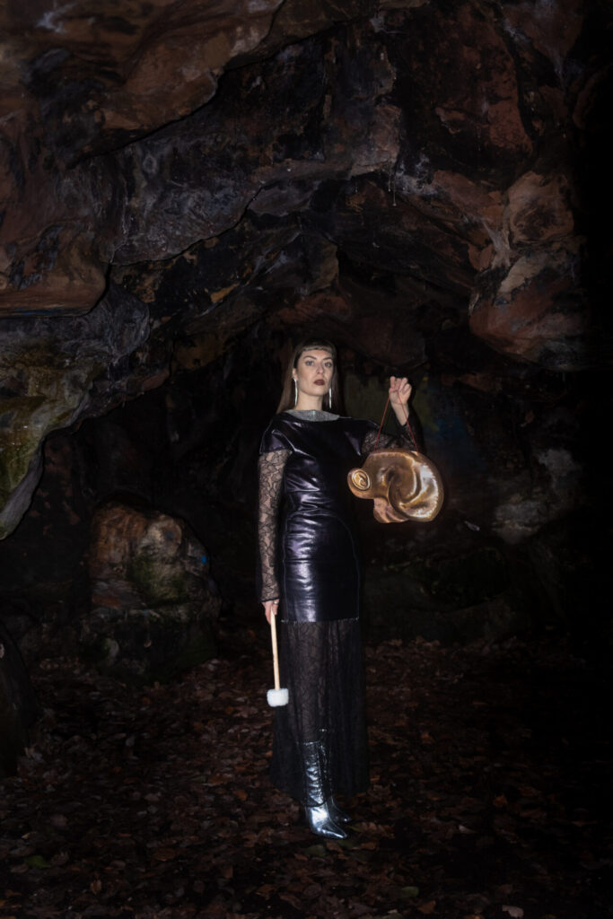 Lola De La Mata holding an ear-shaped gong while stood in a cave